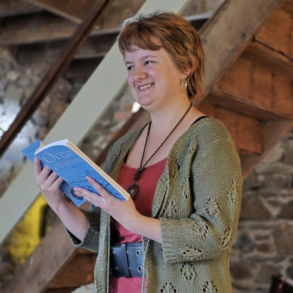 A woman stands indoors, smiling while reading a book titled "Ocean of Emotions." She wears a green cardigan and colorful skirt. The setting is cozy with stone walls and a wooden staircase.