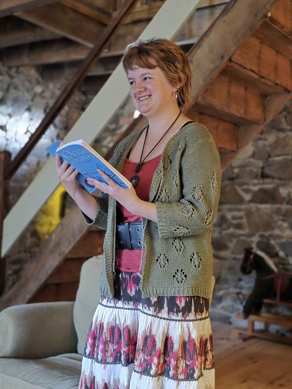 A smiling woman called Lily O. Laverick, stands in a cozy room holding a blue book, written by her. She wears a green knit cardigan and a floral skirt, with a wooden staircase behind her.
