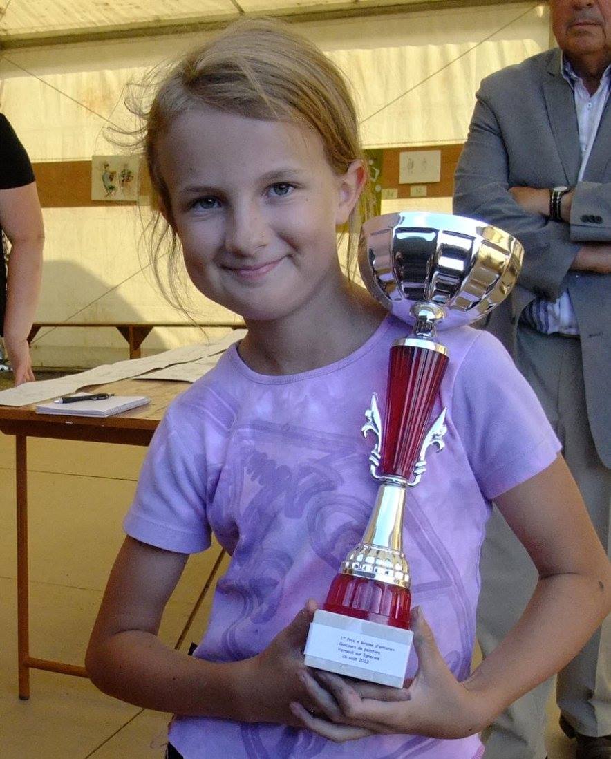 Young girl in a purple shirt proudly holding a large trophy indoors. She smiles confidently, conveying joy and achievement. Adults stand in the background.