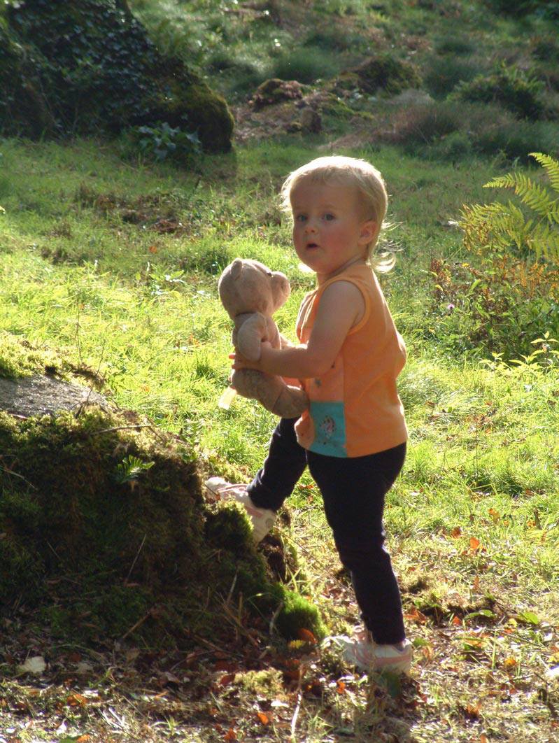 A toddler stands in a sunlit forest, holding a teddy bear. They're wearing an orange top and black pants, looking curiously at their surroundings.